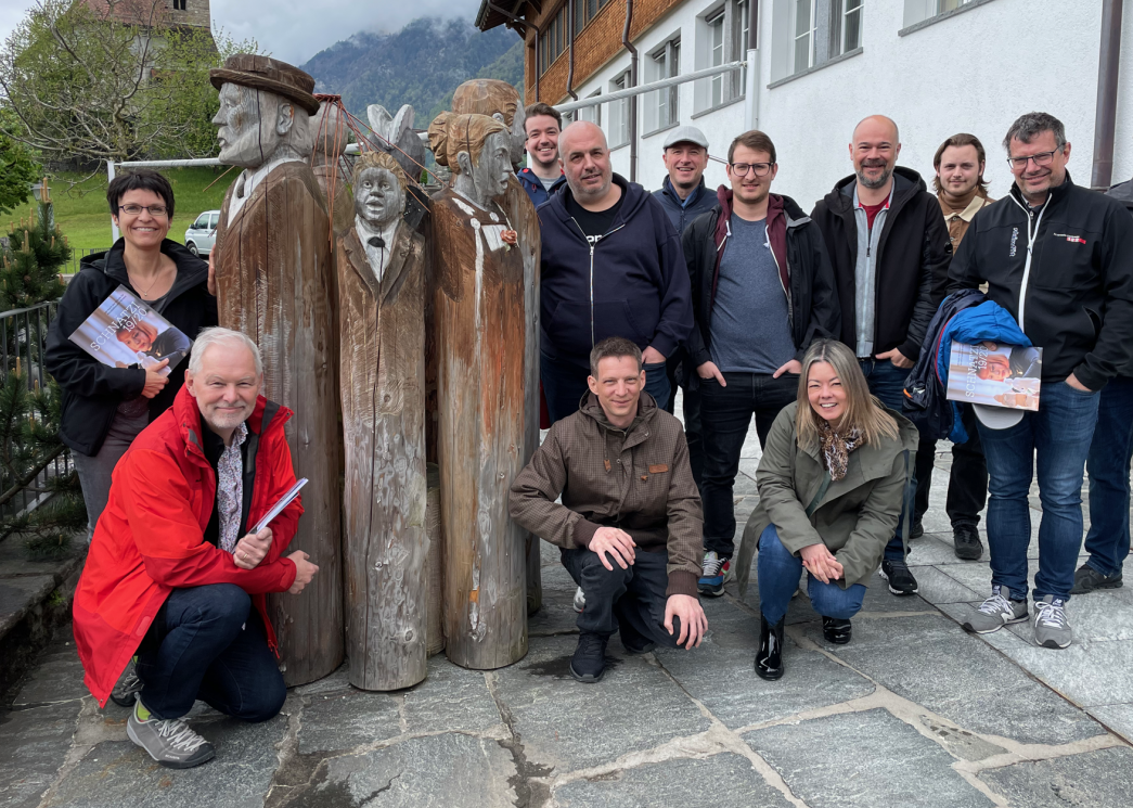Das Topix Team vor der Schnitzlerschule in Brienz. v.l.n.r. Bernadette, Dieter, 7 Holzköpfe, Nicolas, Martin, Daniel, Fabio, Adrian, Aspasia, Sven, Noel, Dietmar. Es fehlt: Reto (am Fotono).